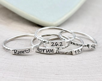 Puede incluir: Un conjunto de cuatro anillos finos de metal plateado, cada uno con una pequeña placa rectangular grabada con texto negro. Las inscripciones incluyen "Run" con un corazón, "26.2", "13.1" y "5k", todos sobre bandas sencillas.
