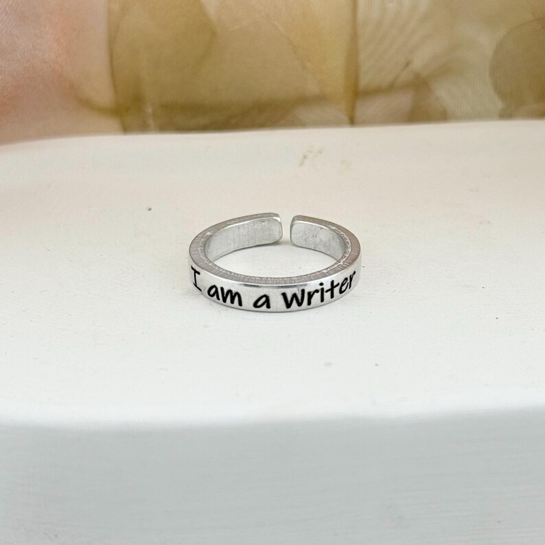 May include: Silver adjustable ring with the text "I am a Writer" engraved on the band. The ring is made of a shiny metal and has a simple, elegant design. The ring is open at the bottom, allowing for size adjustment.