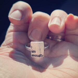May include: A silver ring with a rectangular charm that reads "I am enough".