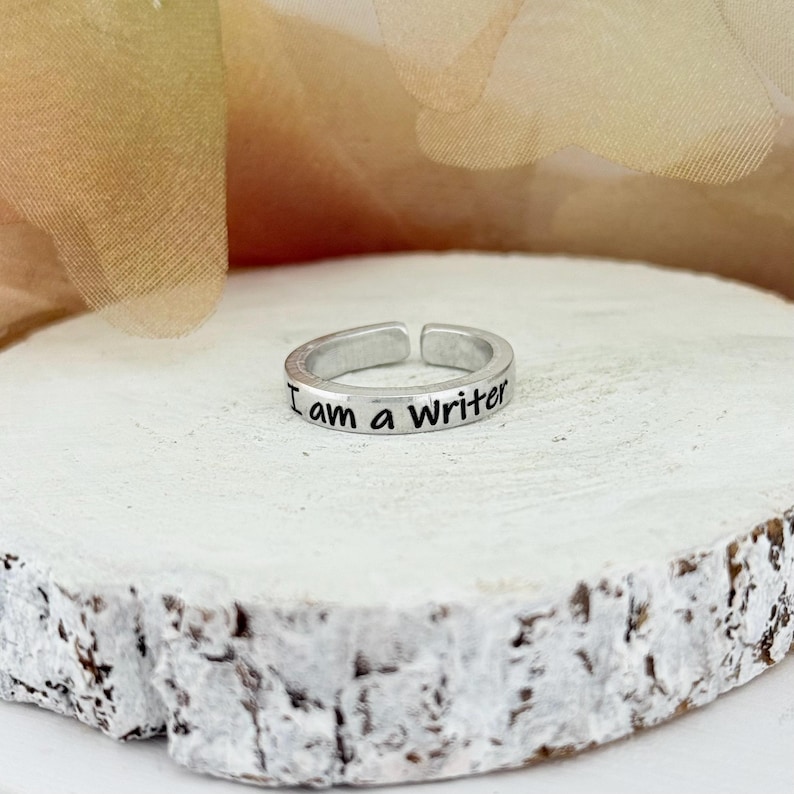 May include: A silver-colored adjustable ring with the inscription "I am a writer" in black lettering. The ring is displayed on a white, textured, wooden surface, with a soft, blurred background.