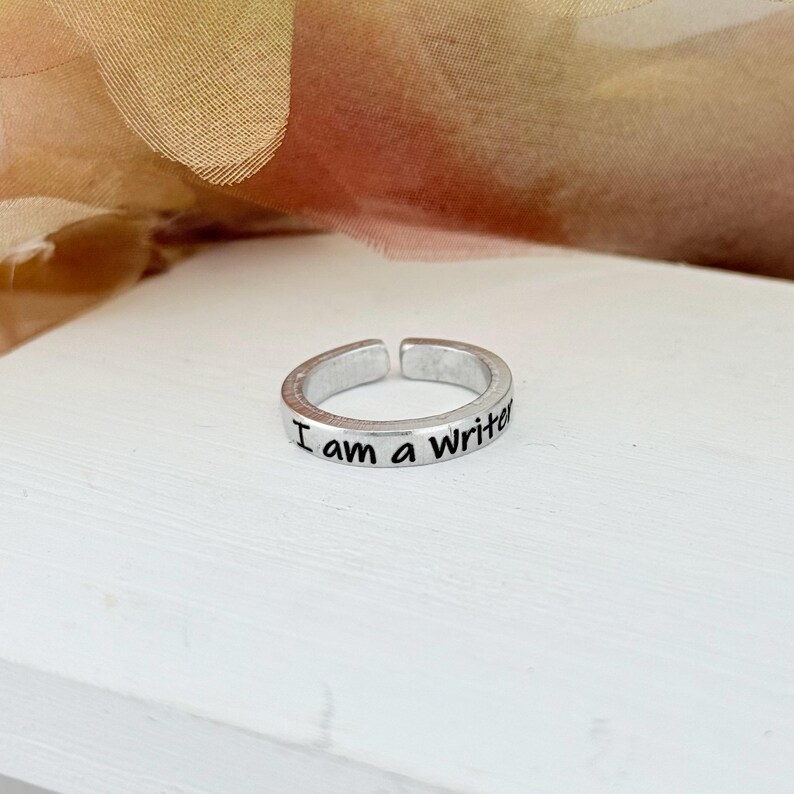 May include: A silver-colored adjustable ring with the words "I am a Writer" engraved on the band. The ring is displayed on a white surface with a soft, neutral-toned background.