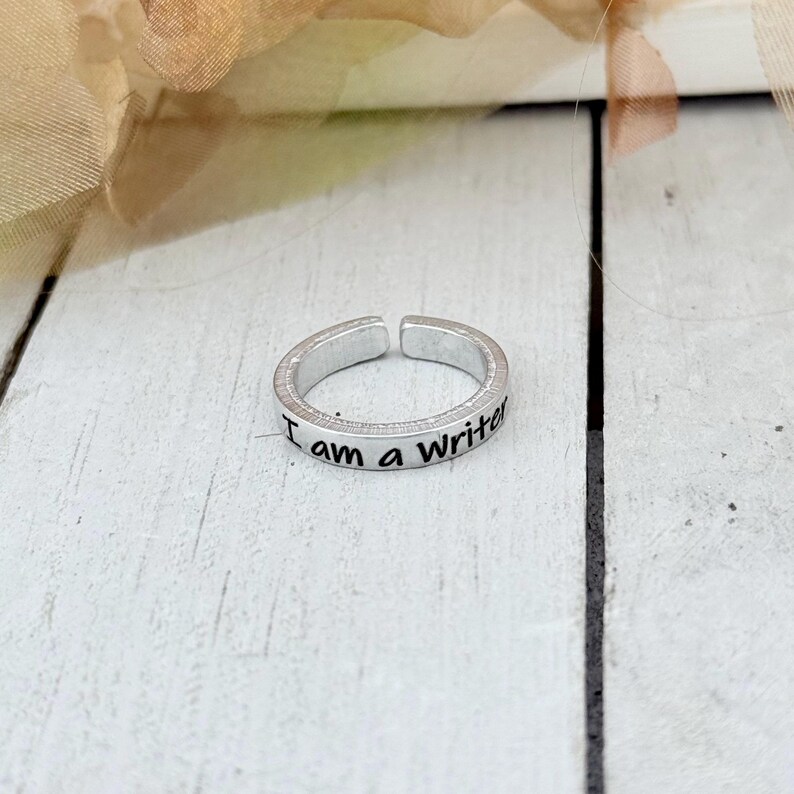 May include: A silver-colored adjustable ring with the words "I am a Writer" engraved on the band. The ring is set against a white wooden surface with a blurred floral background.