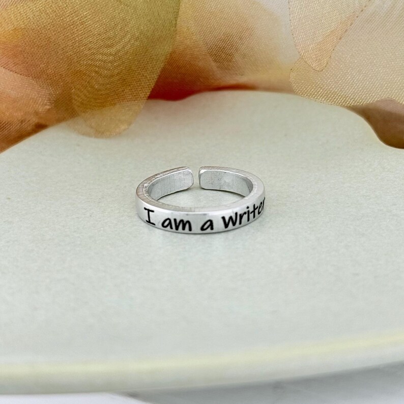May include: A silver-colored adjustable ring with the inscription "I am a Writer" in black lettering. The ring is displayed on a light green surface, with a blurred background of yellow and beige.