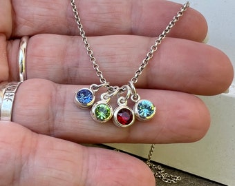May include: A silver chain necklace with five small charms, each featuring a different colored gemstone. The gemstones are blue, green, red, and two shades of blue.