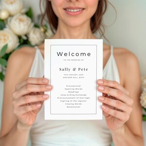 May include: A white wedding program with black text, held by a person. The program reads "Welcome" and lists the names "Sally & Pete" along with the wedding date and location. Floral arrangements are visible in the background.
