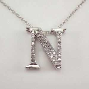 May include: A silver necklace with a diamond-encrusted letter "N" pendant. The pendant hangs from a fine silver chain. The necklace is displayed against a plain white backdrop, emphasizing the jewellery's design and the sparkle of the diamonds.