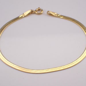 May include: A gold-toned herringbone chain bracelet featuring a flat, flexible link design with a polished finish. The bracelet is secured with a small, circular spring ring clasp at one end and a corresponding metal loop at the other end.