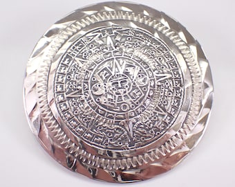 Puede incluir: Un colgante circular de plata con un diseño en relieve detallado del calendario azteca, también conocido como la Piedra del Sol. El colgante tiene un borde texturizado.