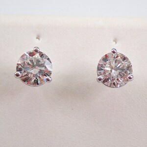 May include: A pair of diamond stud earrings. Each earring features a large, round-cut diamond set in a three-prong white metal setting. The diamonds sparkle with multiple facets, reflecting light. The earrings are displayed against a white background.