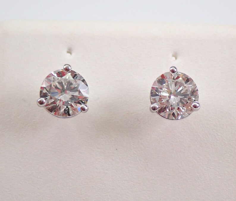 May include: A pair of diamond stud earrings. Each earring features a large, round-cut diamond set in a three-prong white metal setting. The diamonds sparkle with multiple facets, reflecting light. The earrings are displayed against a white background.