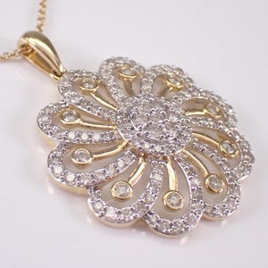 May include: A gold-toned pendant necklace featuring a flower design. The pendant has a central cluster of small, clear stones surrounded by petal-shaped sections, each outlined with more stones. The necklace chain is also gold-toned.