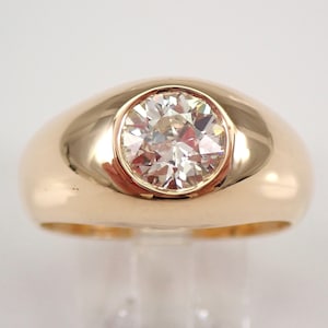 May include: A gold ring with a large, round, clear diamond set in a bezel setting. The ring has a thick band and a domed top.