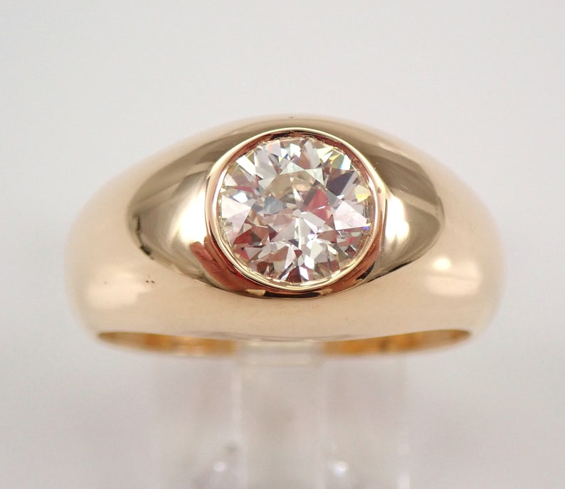 May include: A gold ring with a large, round, clear diamond set in a bezel setting. The ring has a thick band and a domed top.