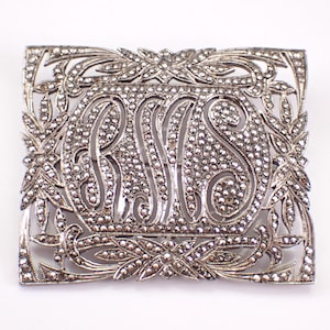 May include: A rectangular, silver-toned brooch with an ornate design. The brooch features the initials "R S S" in the center, surrounded by a decorative border of leaves and small, sparkling embellishments. The overall design is intricate and detailed.