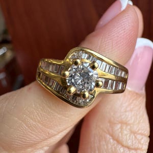 May include: A gold ring featuring a central round-cut clear stone held by six prongs, flanked by rows of channel-set baguette-cut clear stones along the band. The design incorporates a bypass style where the metal curves around the center.