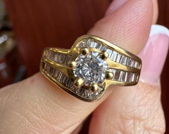 Op de afbeelding: Een gouden ring met een centrale ronde heldere steen vastgezet met zes klauwen, geflankeerd door rijen in kanaal gezette heldere baguette-geslepen stenen langs de band. Het ontwerp heeft een bypass-stijl waarbij het metaal buigt.