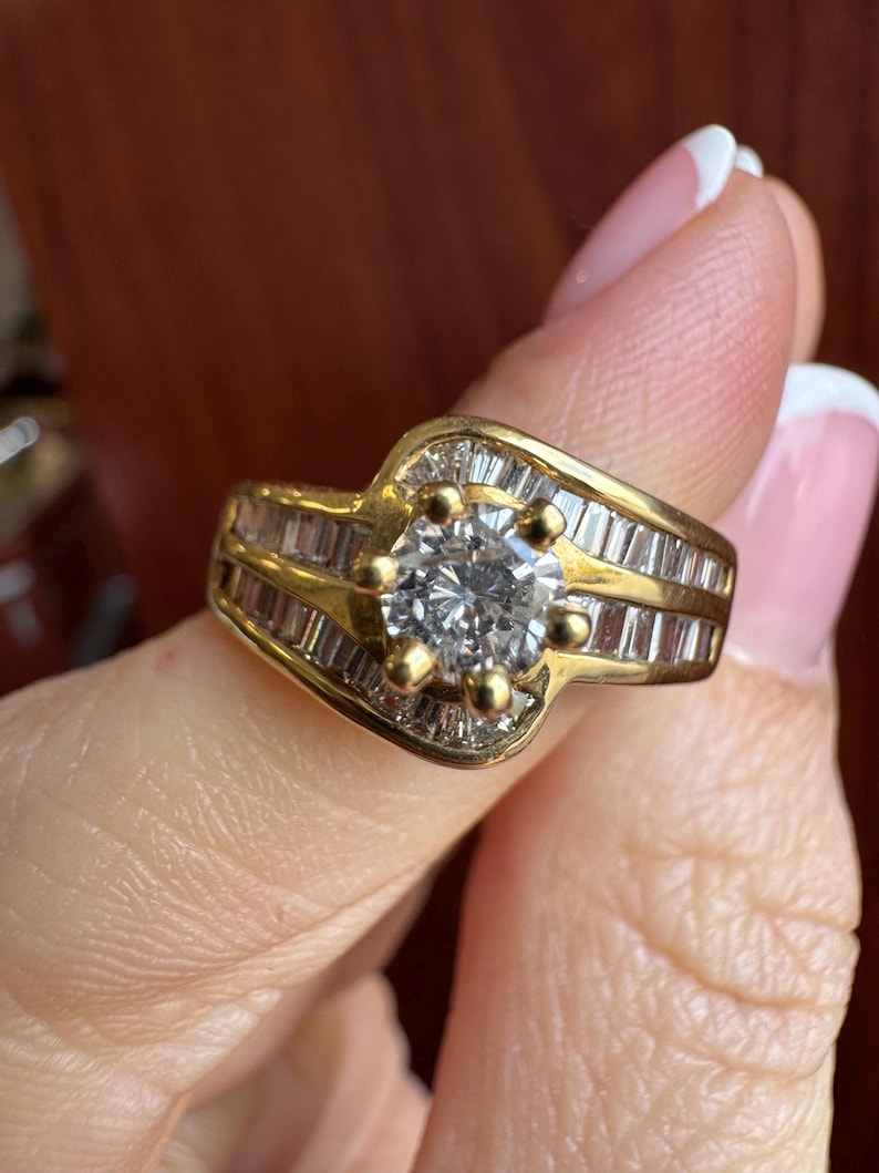 May include: A gold ring featuring a central round-cut clear stone held by six prongs, flanked by rows of channel-set baguette-cut clear stones along the band. The design incorporates a bypass style where the metal curves around the center.