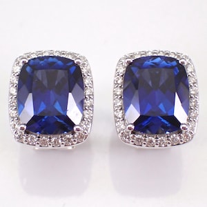 Pode incluir: Um par de brincos de botão quadrados com pedras preciosas azuis. Cada brinco apresenta uma grande pedra central azul escura, rodeada por um halo de pedras preciosas menores e claras. Os brincos são cravejados em metal prateado.