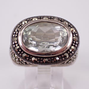 May include: A silver ring with an oval, light green gemstone. The gemstone is surrounded by a border of small, dark, round stones. The ring has a detailed, textured band.