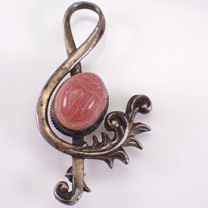 May include: A silver-toned brooch in the shape of a treble clef, featuring a pink oval stone at its center. The brooch has an ornate, scrolling design at the bottom. The stone has a textured appearance.