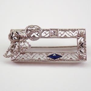 Può includere: Una spilla rettangolare color argento con un intricato telaio in filigrana traforata. Il design include un fiocco decorativo sul lato sinistro, una singola pietra rotonda trasparente in alto e una pietra blu scuro taglio marquise in basso.