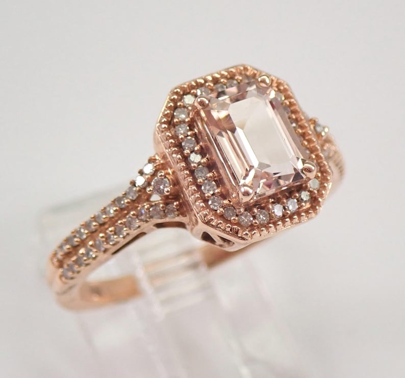 May include: A rose gold engagement ring with a large, rectangular, light pink morganite gemstone surrounded by a halo of smaller diamonds. The band is also set with smaller diamonds.