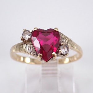 Op de afbeelding: Een goudkleurige ring met een hartvormige, dieprode edelsteen als middelpunt. Twee kleinere, heldere edelstenen flankeren het hart. De band heeft een textuur.