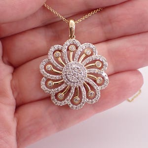 May include: A gold pendant necklace featuring a flower design. The pendant has a central cluster of small, round diamonds surrounded by petals of diamonds and gold. The necklace is held in a hand.