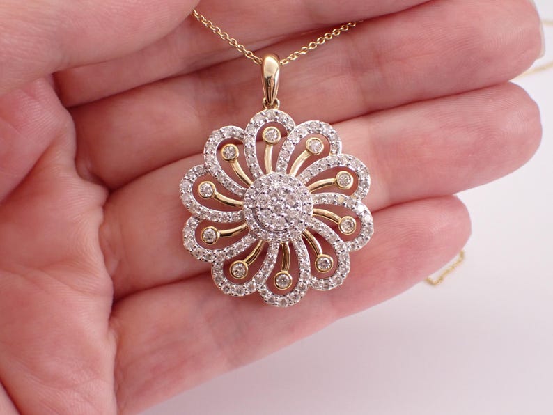 May include: A gold pendant necklace featuring a flower design. The pendant has a central cluster of small, round diamonds surrounded by petals of diamonds and gold. The necklace is held in a hand.