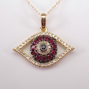 May include: Gold-tone pendant necklace with an eye-shaped charm. The charm is decorated with red and white gemstones, and a black central stone. The pendant hangs from a fine gold chain.