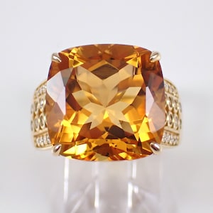 May include: A gold ring with a large, cushion-cut topaz gemstone surrounded by smaller diamonds. The ring has a detailed band with a repeating pattern of diamonds.