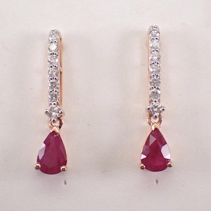 May include: A pair of elegant drop earrings. Each earring features a teardrop-shaped, deep red ruby gemstone suspended from a gold setting. The top portion of each earring is a slender bar adorned with small, sparkling white gemstones.