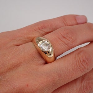 May include: A gold ring with a large, round, clear gemstone set in a bezel setting. The ring is on a person's finger.