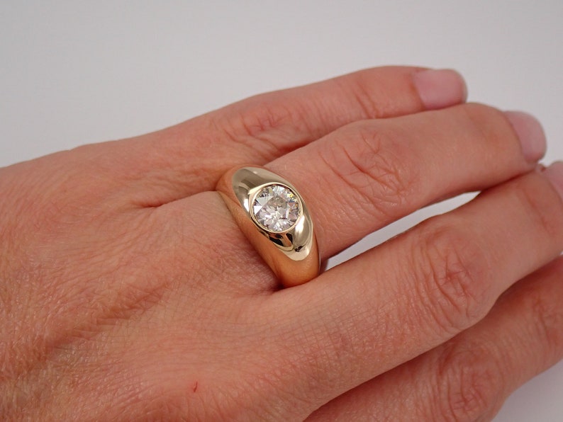 May include: A gold ring with a large, round, clear gemstone set in a bezel setting. The ring is on a person's finger.