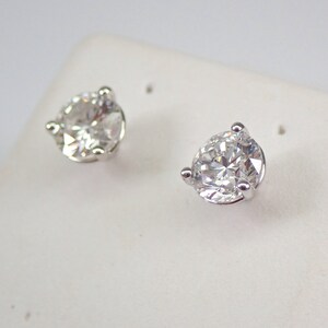 May include: A pair of clear, round-cut diamond stud earrings. Each earring features a single, sparkling diamond held in a silver-colored, three-prong setting. The earrings are displayed on a white surface.