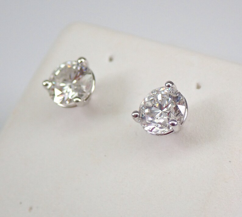 May include: A pair of clear, round-cut diamond stud earrings. Each earring features a single, sparkling diamond held in a silver-colored, three-prong setting. The earrings are displayed on a white surface.