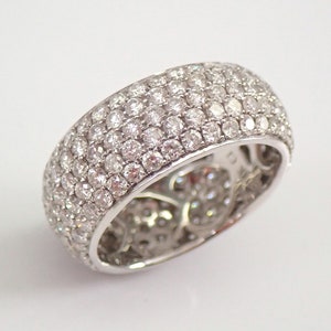 Op de afbeelding: Een brede, zilverkleurige ring die dicht bezet is met talrijke kleine, ronde, heldere edelstenen. Het oppervlak van de ring is volledig bedekt met de sprankelende stenen, wat een briljant, gestructureerd uiterlijk creëert. De ring staat tegen een effen witte achtergrond.