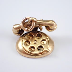 Könnte beinhalten: Ein goldfarbener Vintage-Telefonanhänger. Der Anhänger verfügt über ein Wählscheibe und einen Hörer mit einer Schlaufe zum Befestigen an einer Halskette oder einem Armband. Der Telefonanhänger ist ein klassisches Design, ideal für Schmuck.