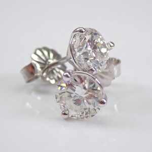 May include: A pair of diamond stud earrings. Each earring features a large, round, brilliant-cut diamond set in a white metal prong setting. The earrings are displayed on a white surface, with the earring backs visible.