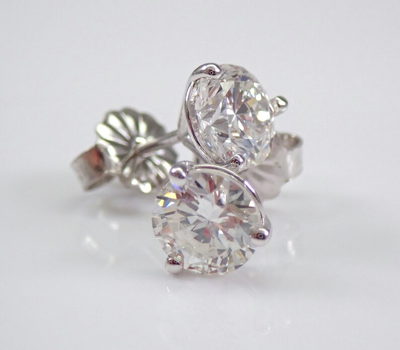 May include: A pair of diamond stud earrings. Each earring features a large, round, brilliant-cut diamond set in a white metal prong setting. The earrings are displayed on a white surface, with the earring backs visible.