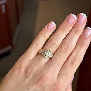 May include: A gold ring featuring a central round brilliant-cut diamond flanked by rows of channel-set baguette diamonds. The band has a bypass design, with the metal curving around the center stone to create a layered, flowing appearance.