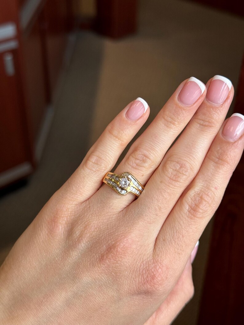May include: A gold ring featuring a central round brilliant-cut diamond flanked by rows of channel-set baguette diamonds. The band has a bypass design, with the metal curving around the center stone to create a layered, flowing appearance.