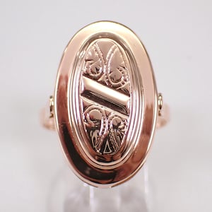 May include: Rose gold oval ring with an engraved design. The ring features an oval face with an ornate, symmetrical pattern. The band is a simple, polished rose gold. The ring is set against a white background.