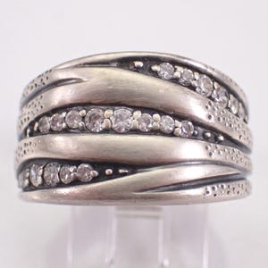 Puede incluir: Un anillo plateado con un diseño texturizado y en capas. El anillo presenta múltiples bandas con piedras preciosas claras incrustadas. Las bandas tienen un patrón ondulado y están acentuadas con pequeños detalles punteados.