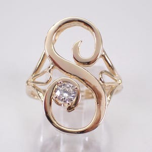May include: A gold ring with a swirling design, featuring a central, clear gemstone. The ring's design incorporates an open, cursive letter "S" shape, with the gemstone set within the lower curve. The ring is displayed on a clear stand.
