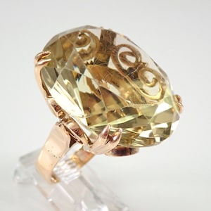 May include: A gold ring with a large, faceted, oval-shaped, light yellow gemstone. The setting is intricate and features a delicate, swirling design.