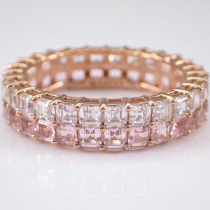 May include: A rose gold eternity band ring with two rows of square-cut gemstones. The top row features clear gemstones, while the bottom row features pink gemstones.