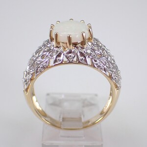 May include: A gold ring featuring a large, oval, white opal gemstone. The band has a detailed design with small, clear gemstones and a filigree pattern. The ring is displayed on a clear stand, highlighting its intricate details.