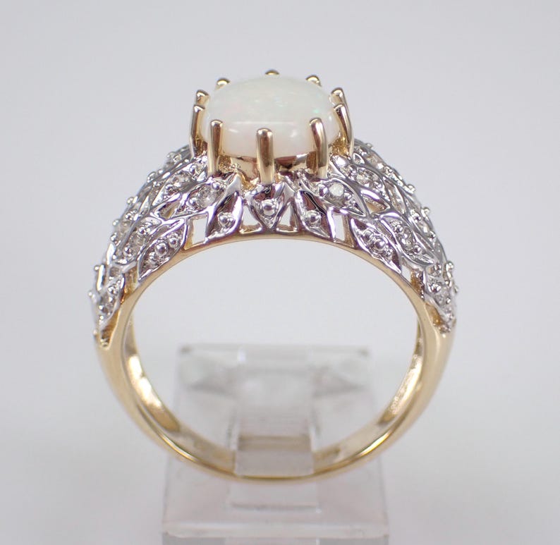 May include: A gold ring featuring a large, oval, white opal gemstone. The band has a detailed design with small, clear gemstones and a filigree pattern. The ring is displayed on a clear stand, highlighting its intricate details.