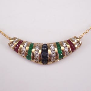 Puede incluir: Un collar de oro con un colgante curvo. El colgante presenta filas alternas de piedras preciosas rojas, verdes, blancas y azul oscuro. La cadena del collar también es de oro.